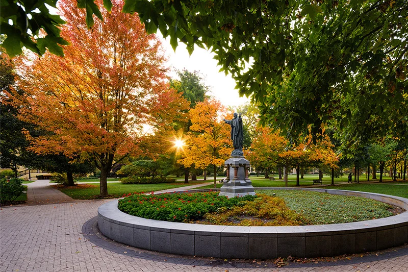 Christ the Teacher statue on Notre Dame's God Quad, surrounded by vibrant orange and yellow autumn trees under a golden sunburst.