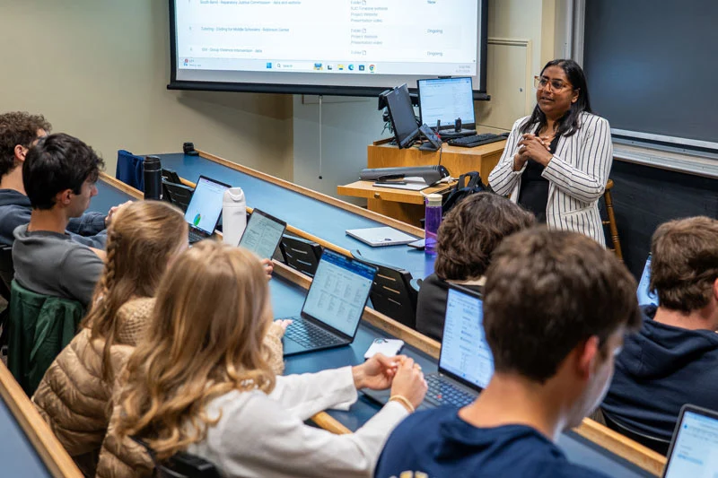 A woman with dark hair stands at the front of a college classroom.