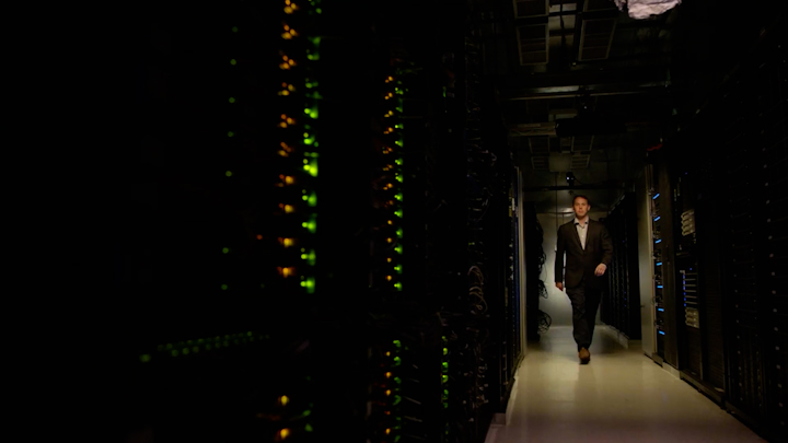 A man in a dark suit walks through a dimly lit data center corridor lined with server racks glowing with green and amber indicator lights.