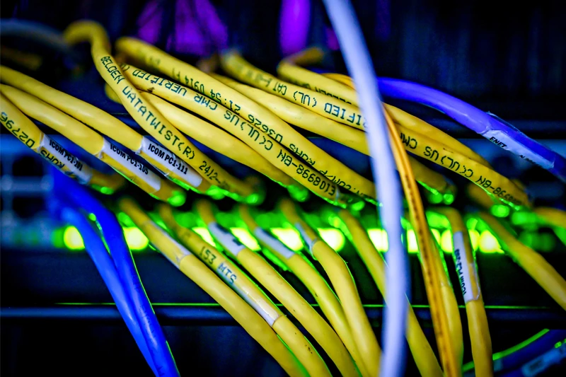 A tangled bundle of yellow and blue ethernet cables is illuminated by the green lights of network equipment.