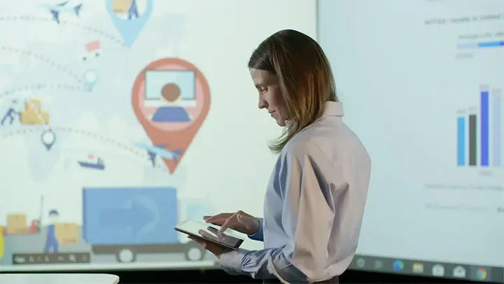 Woman in a light blue shirt taps a tablet, facing a large screen displaying colorful logistics maps and data charts.