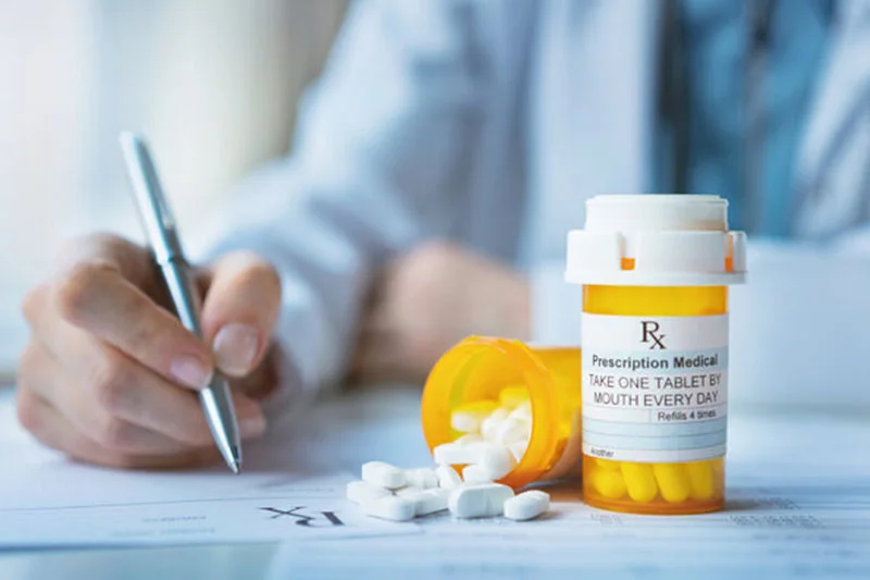 A hand in a lab coat writes on a prescription pad. Nearby, white pills spill from an overturned bottle; another bottle contains yellow pills.