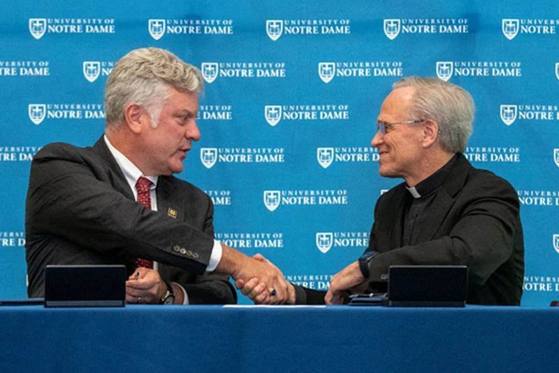 Father John I. Jenkins, C.S.C. shakes hands with a gray-haired man in a dark suit against a blue Notre Dame backdrop.