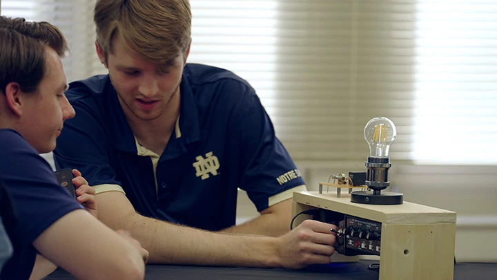 Two Notre Dame students in navy polos work on an electronic device with a light bulb, one adjusting a knob.