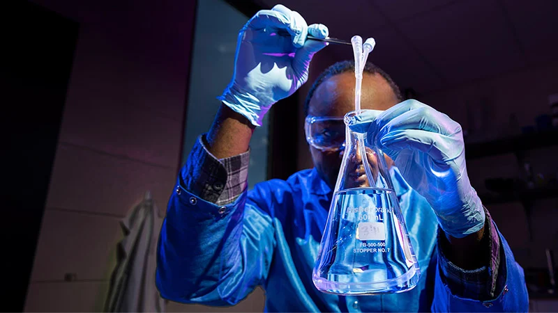 A researcher wearing safety goggles, a lab coat, and blue nitrile gloves uses tweezers to lower tubing into a glass flask filled with clear liquid.