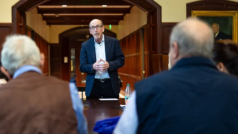 A man in a suit jacket and glasses stands speaking at the head of a conference table, addressing a group of seated people viewed from behind in a room with wood paneling and arched doorways.